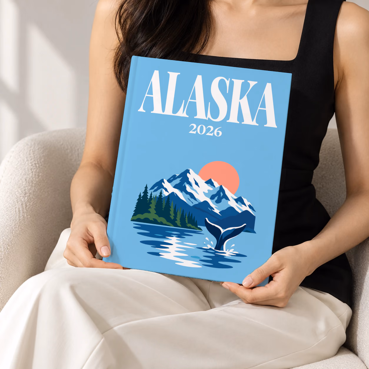 Alaska hardcover photo book with the illustrated cover of a whale tail before snowy mountains and pine forest on sky blue, held upright in two hands by a woman in a black top, sitting in a cream boucle armchair