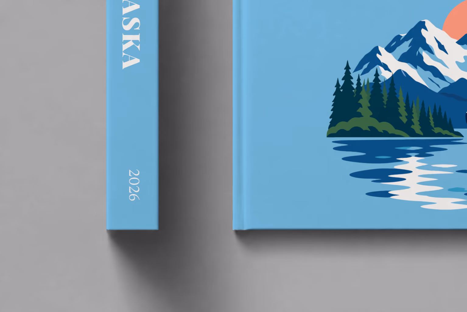 Alaska hardcover photo book with the illustrated cover of a whale tail before snowy mountains and pine forest on sky blue, shown close up beside its spine on a pale grey studio surface
