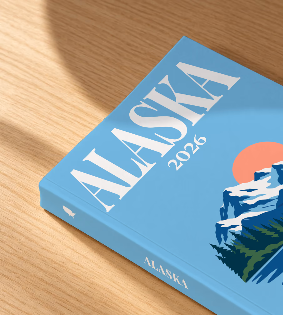 Alaska softcover photo book with the illustrated cover of a whale tail before snowy mountains and pine forest on sky blue, lying on an oak desk in dappled afternoon sunlight