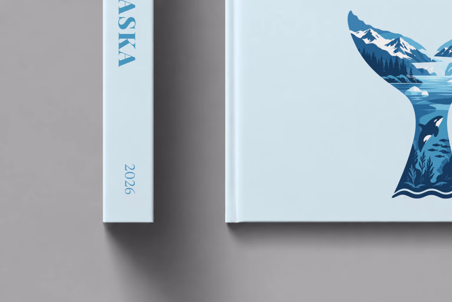 Alaska hardcover photo book with the illustrated cover of a whale tail filled with glaciers, mountains and an orca on pale ice blue, shown close up beside its spine on a pale grey studio surface