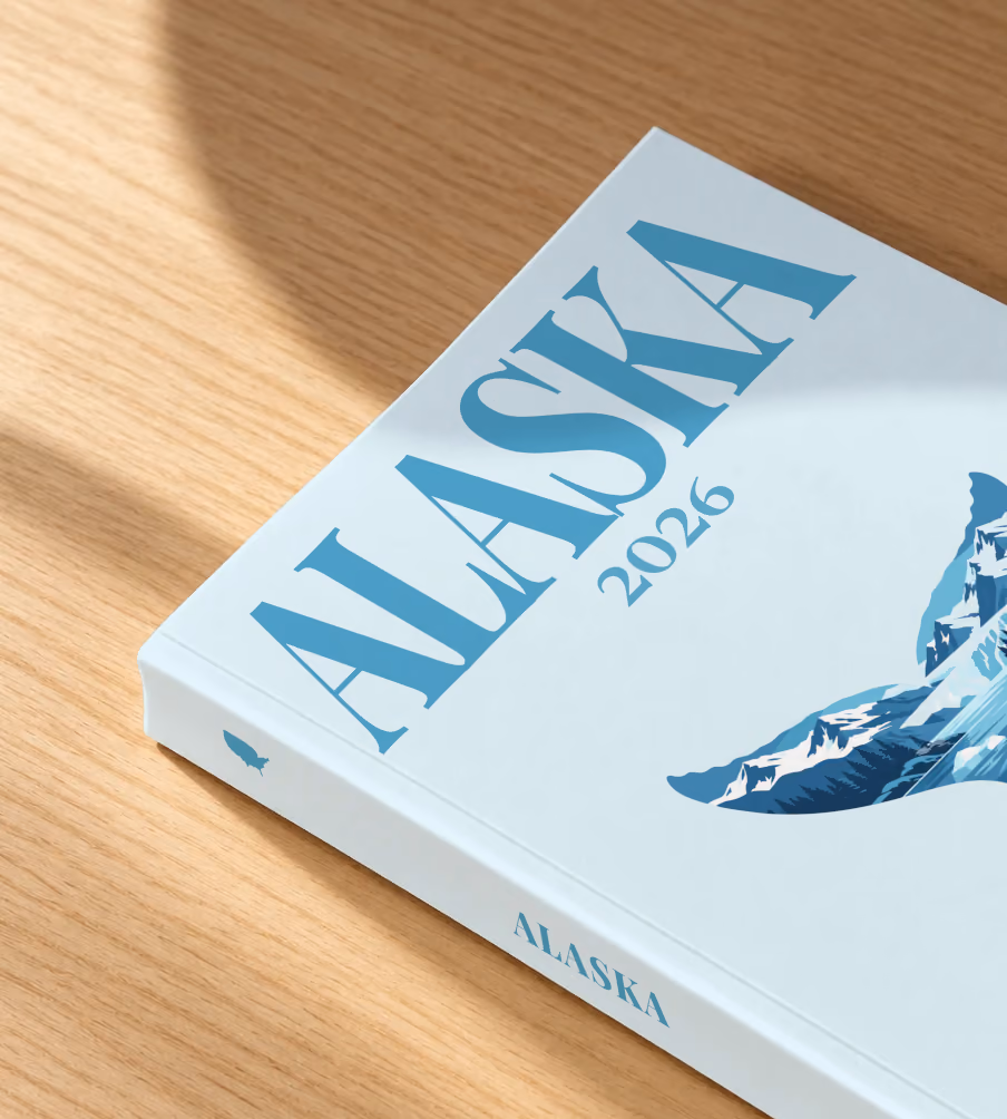 Alaska softcover photo book with the illustrated cover of a whale tail filled with glaciers, mountains and an orca on pale ice blue, lying on an oak desk in dappled afternoon sunlight