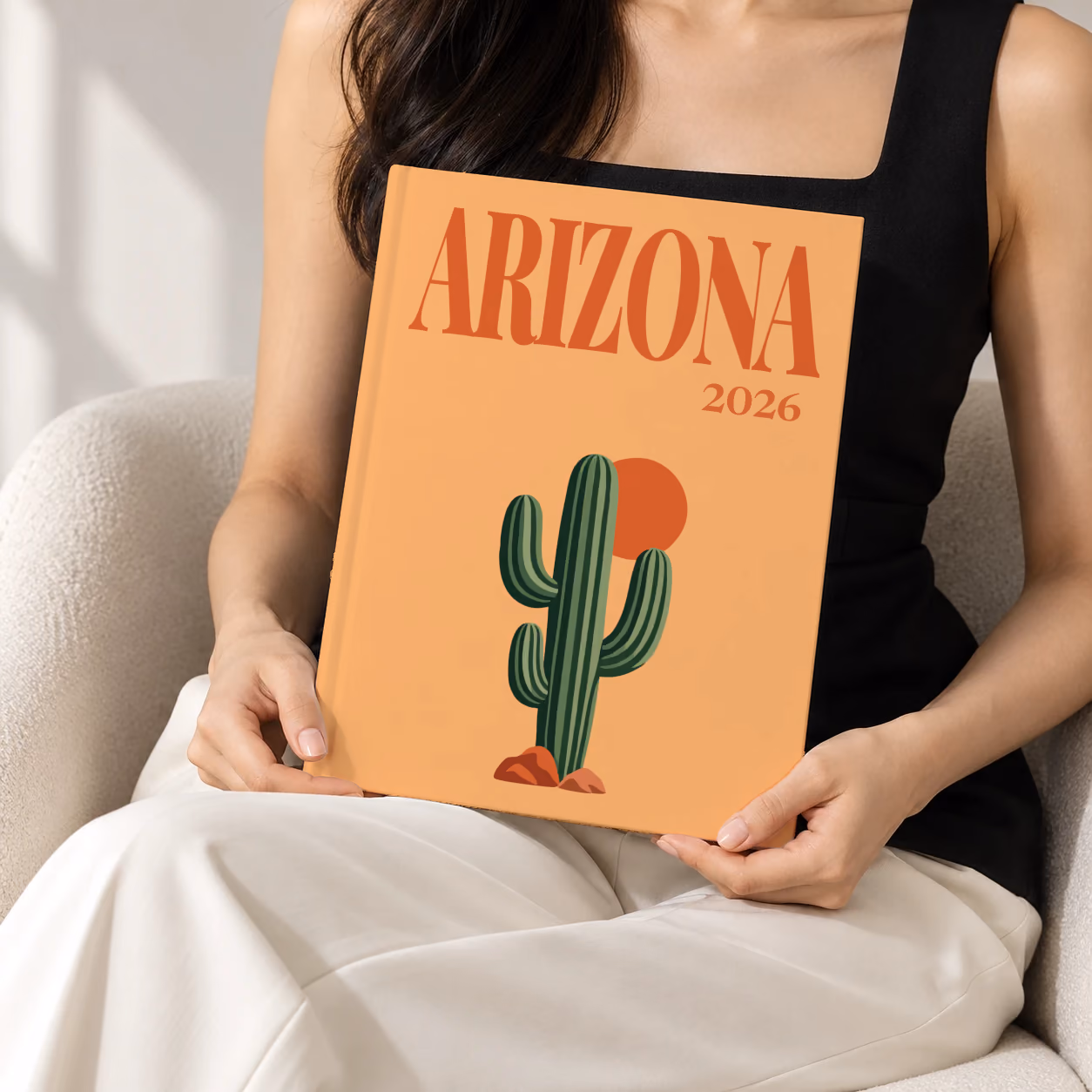 Arizona hardcover photo book with the illustrated cover of a saguaro cactus and a setting sun on apricot, held upright in two hands by a woman in a black top, sitting in a cream boucle armchair