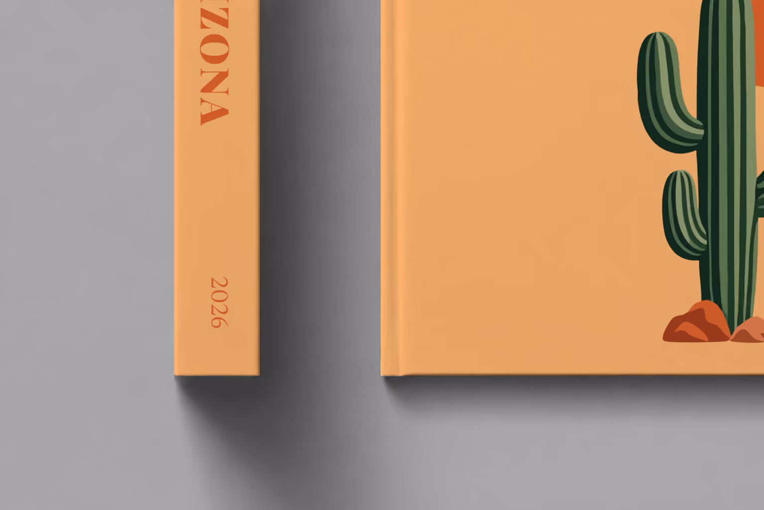 Arizona hardcover photo book with the illustrated cover of a saguaro cactus and a setting sun on apricot, shown close up beside its spine on a pale grey studio surface