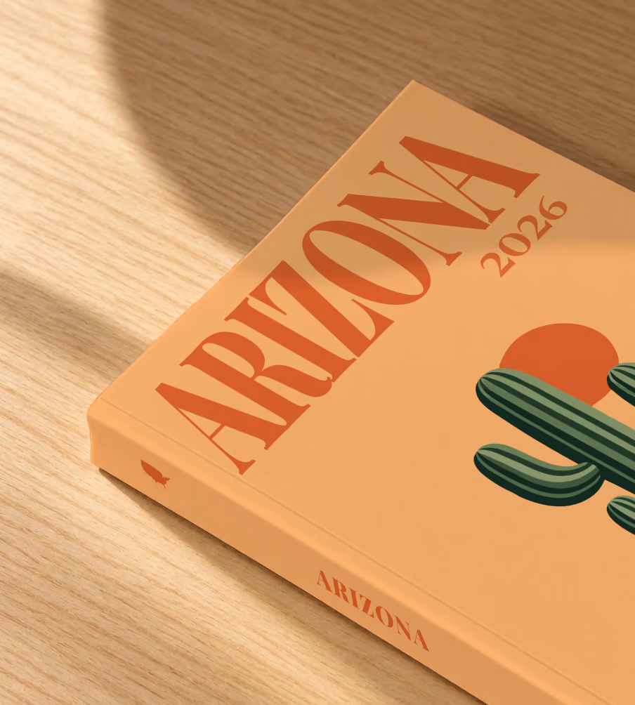 Arizona softcover photo book with the illustrated cover of a saguaro cactus and a setting sun on apricot, lying on an oak desk in dappled afternoon sunlight