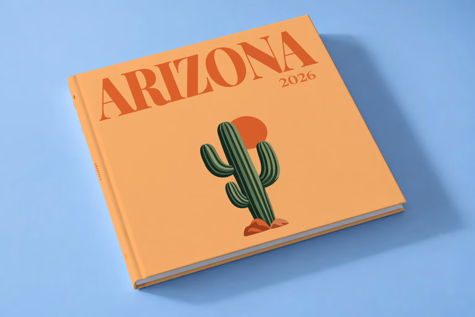 Arizona square hardcover photo book with the illustrated cover of a saguaro cactus and a setting sun on apricot, lying on a pale blue studio backdrop