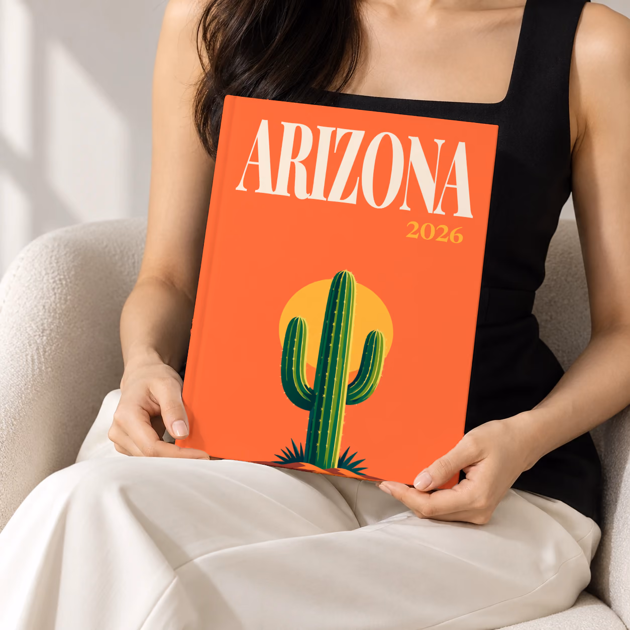 Arizona hardcover photo book with the illustrated cover of a saguaro cactus against a yellow sun on orange, held upright in two hands by a woman in a black top, sitting in a cream boucle armchair