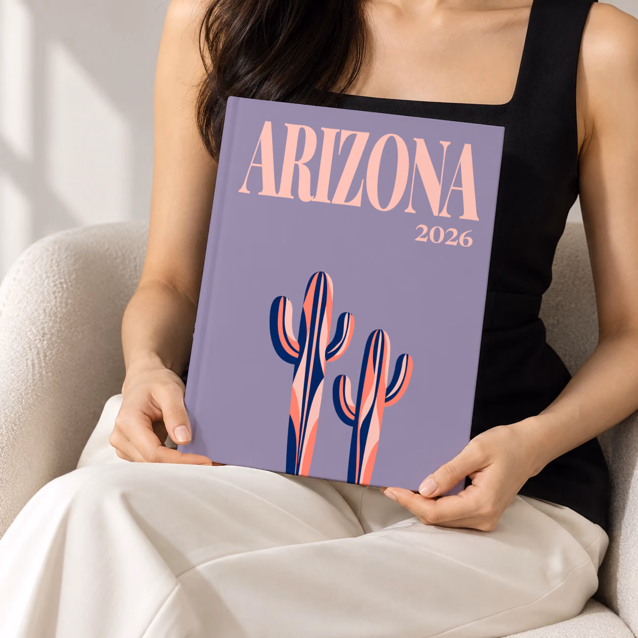 Arizona hardcover photo book with the illustrated cover of two stylised saguaro cacti on dusty purple, held upright in two hands by a woman in a black top, sitting in a cream boucle armchair
