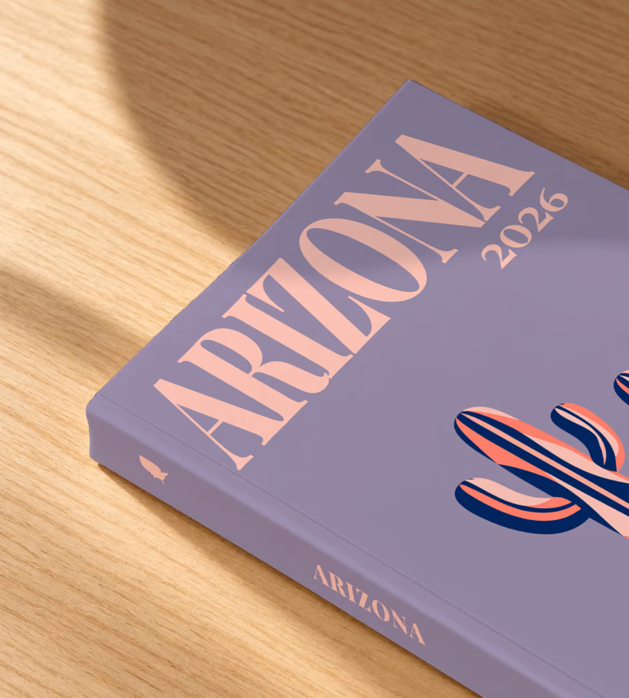 Arizona softcover photo book with the illustrated cover of two stylised saguaro cacti on dusty purple, lying on an oak desk in dappled afternoon sunlight