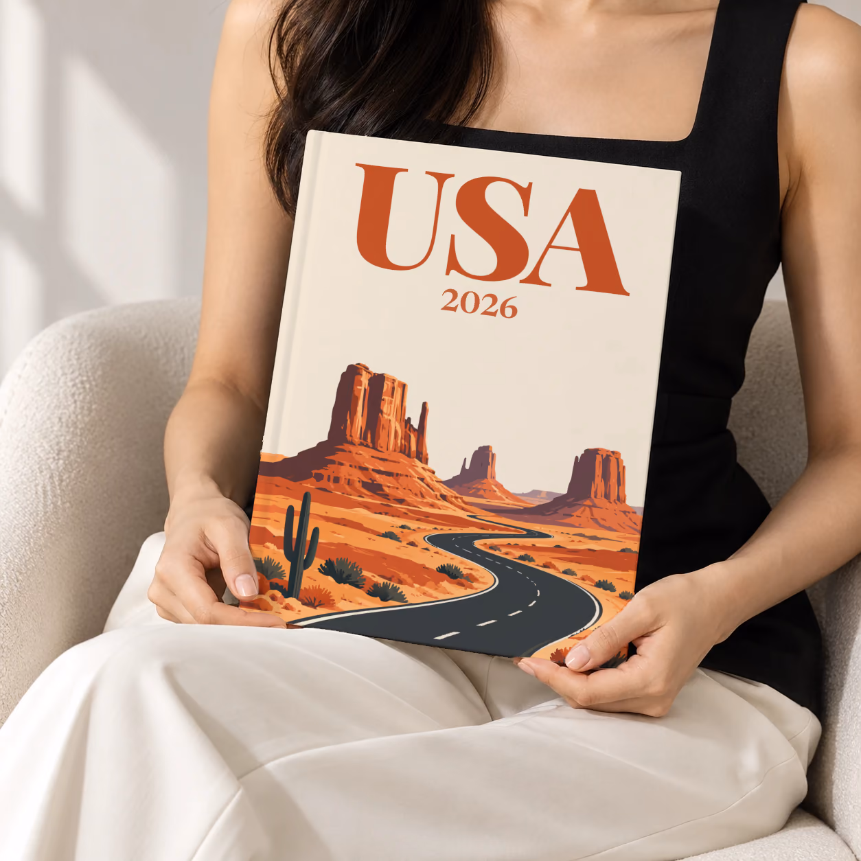 Arizona hardcover photo book with the USA illustrated cover of the road through the red buttes of Monument Valley on sand, held upright in two hands by a woman in a black top, sitting in a cream boucle armchair