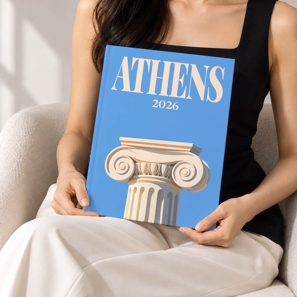 Athens hardcover photo book with the illustrated cover of an Ionic capital on blue, held upright in two hands by a woman in a black top, sitting in a cream boucle armchair