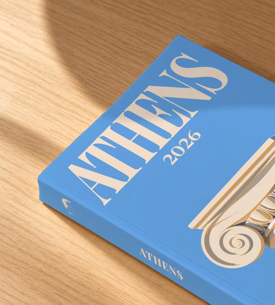 Athens softcover photo book with the illustrated cover of an Ionic capital on blue, lying on an oak desk in dappled afternoon sunlight