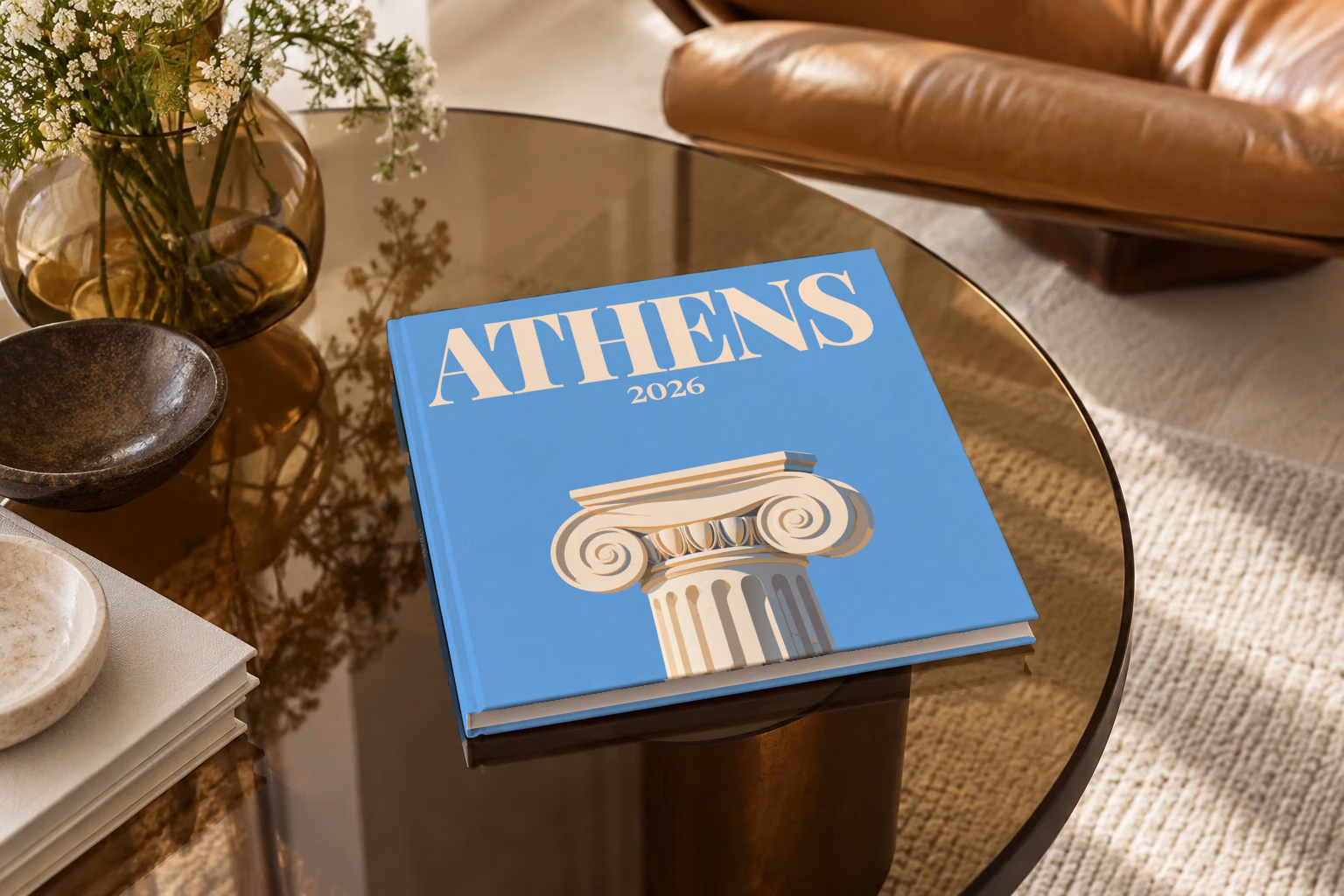 Athens square hardcover photo book with the illustrated cover of an Ionic capital on blue, on a smoked glass coffee table beside a leather armchair in warm afternoon light