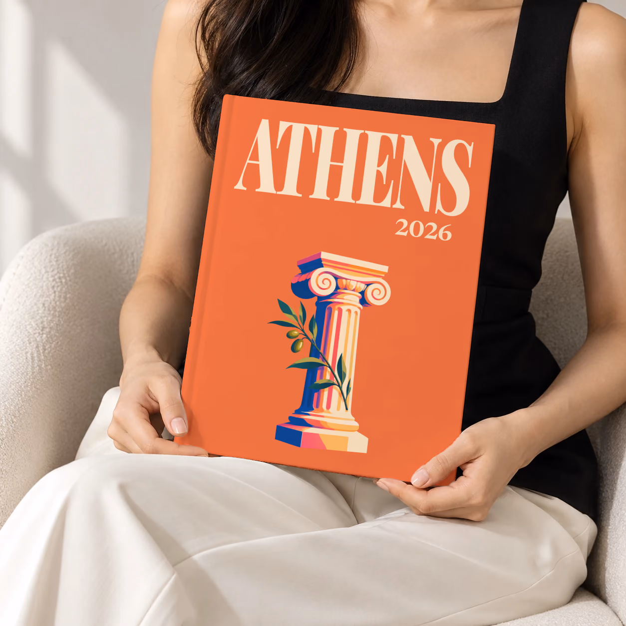 Athens hardcover photo book with the illustrated cover of an Ionic column and an olive sprig on orange, held upright in two hands by a woman in a black top, sitting in a cream boucle armchair