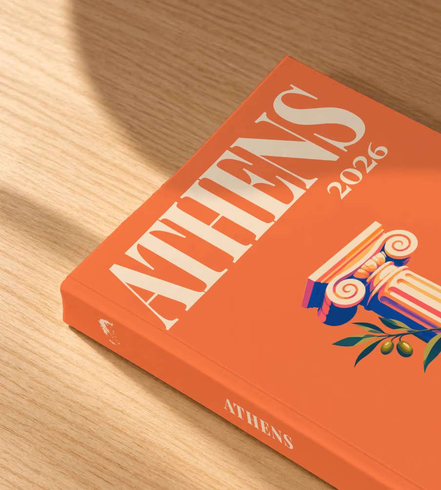 Athens softcover photo book with the illustrated cover of an Ionic column and an olive sprig on orange, lying on an oak desk in dappled afternoon sunlight