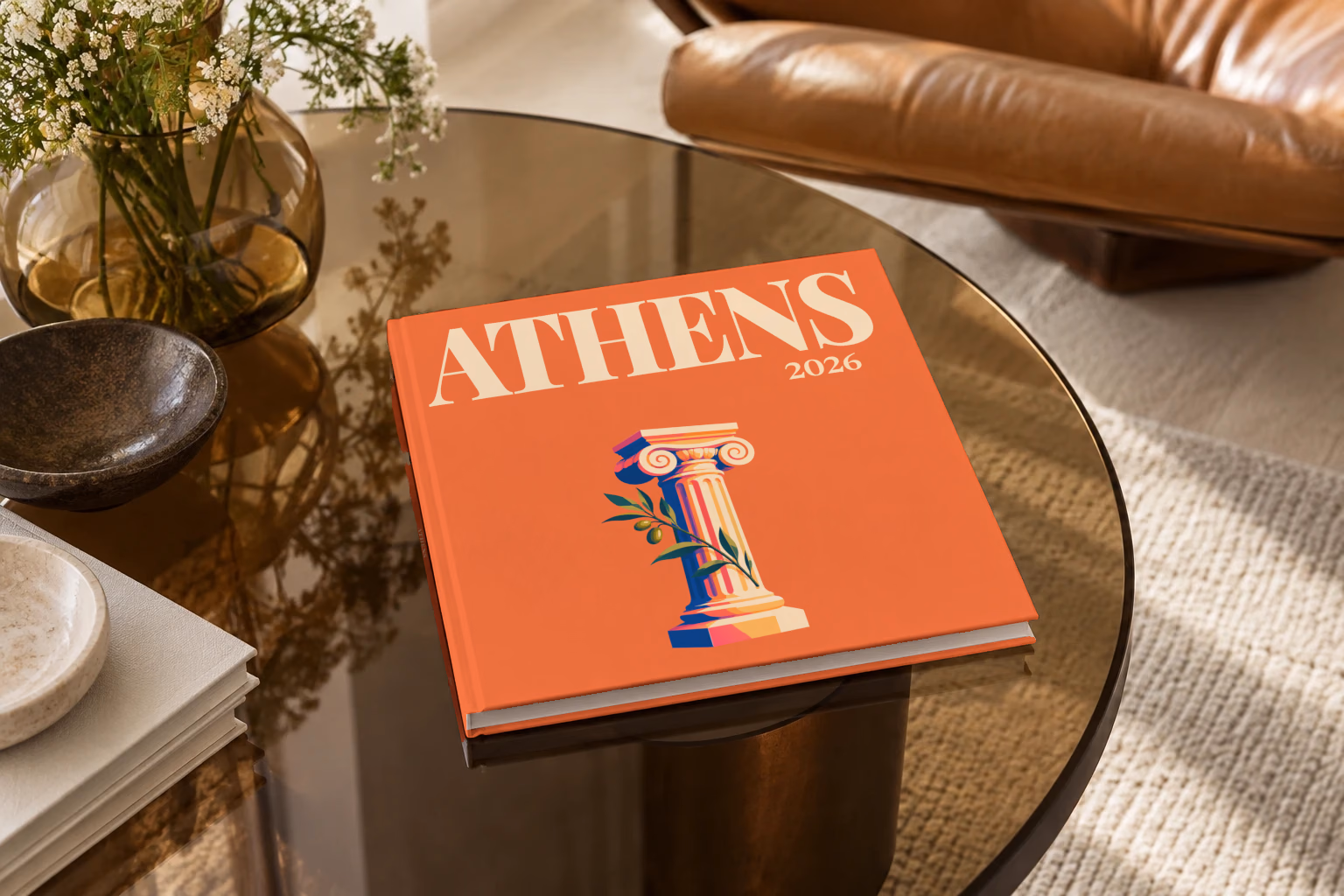 Athens square hardcover photo book with the illustrated cover of an Ionic column and an olive sprig on orange, on a smoked glass coffee table beside a leather armchair in warm afternoon light