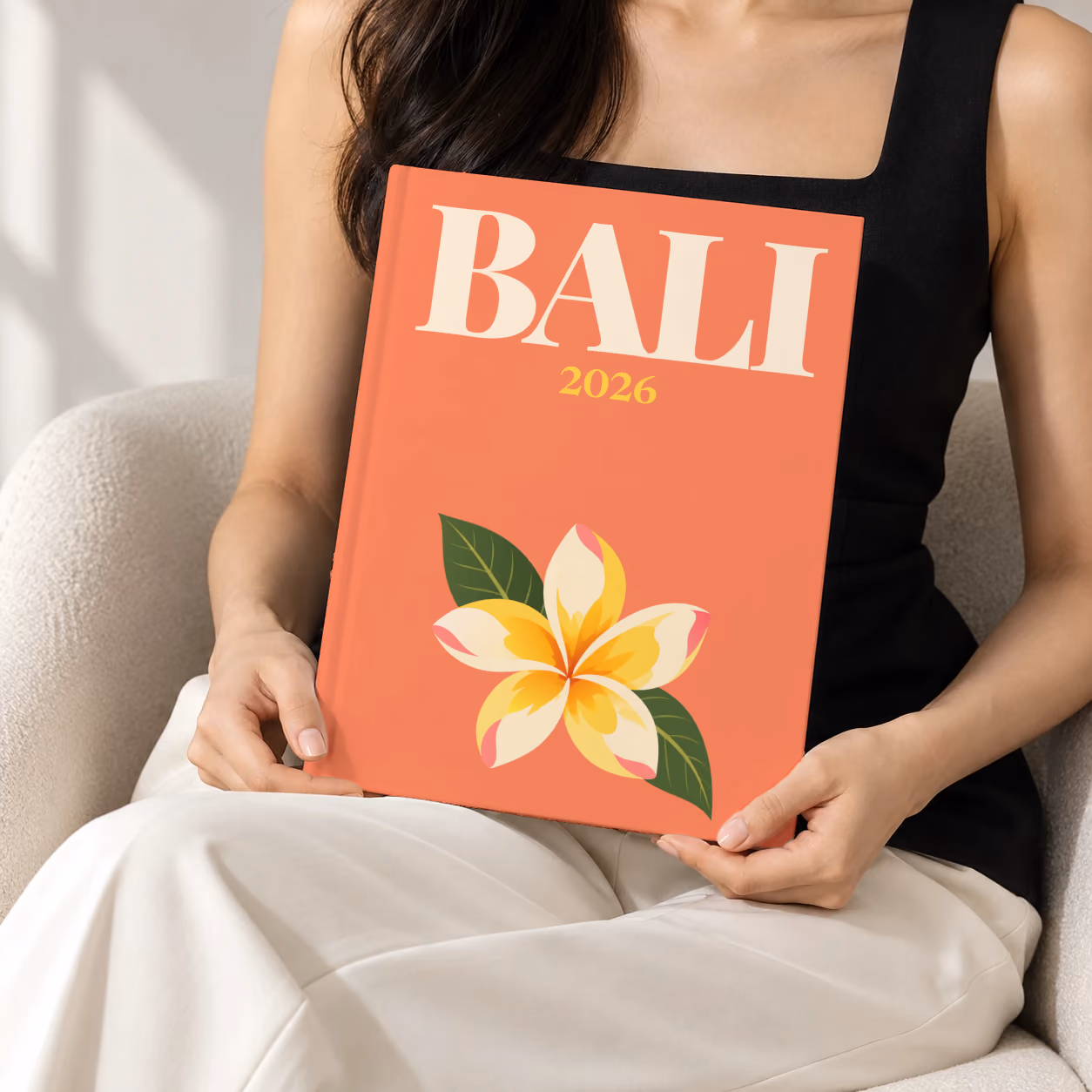 Bali hardcover photo book with the illustrated cover of a yellow frangipani flower on coral, held upright in two hands by a woman in a black top, sitting in a cream boucle armchair