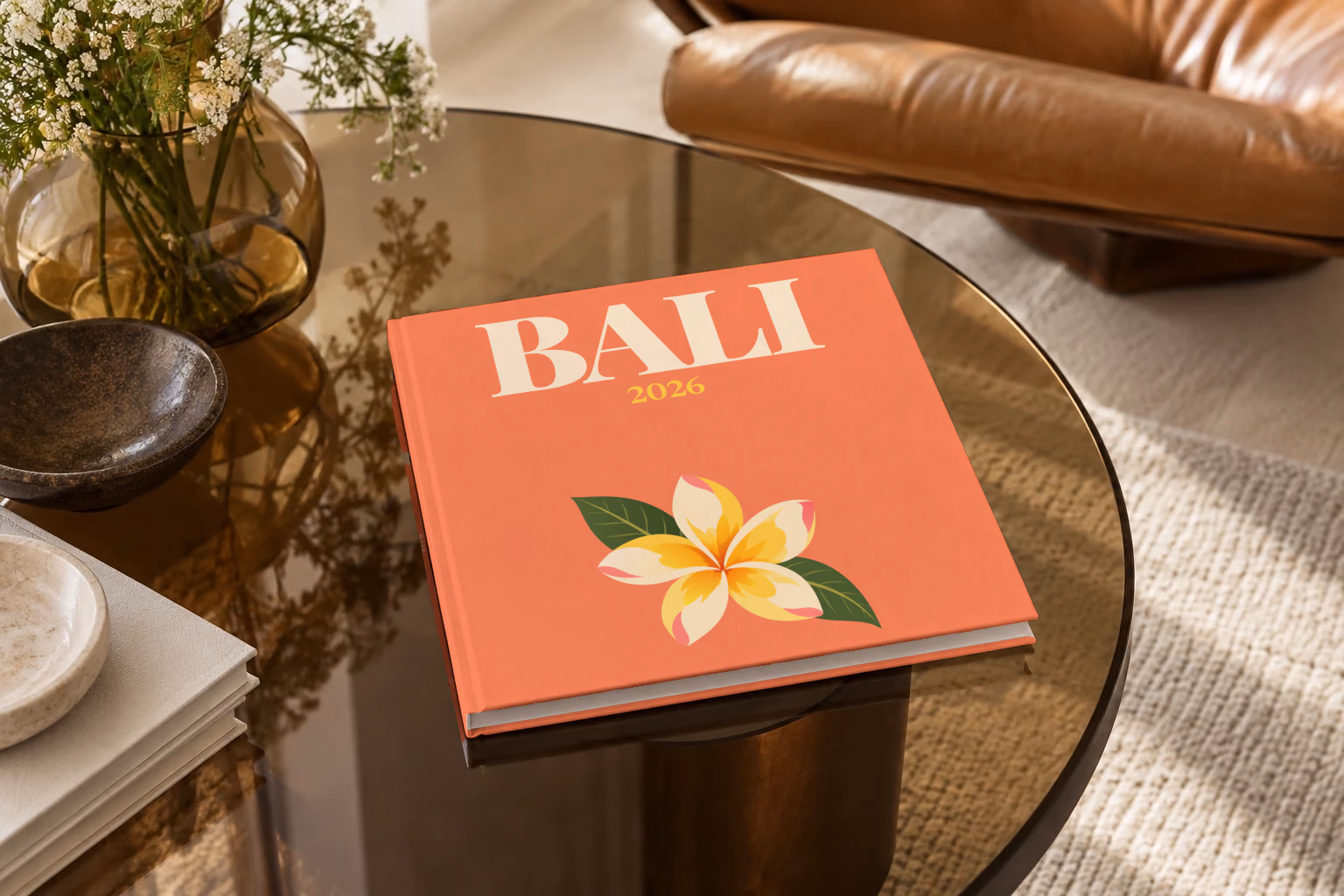 Bali square hardcover photo book with the illustrated cover of a yellow frangipani flower on coral, on a smoked glass coffee table beside a leather armchair in warm afternoon light