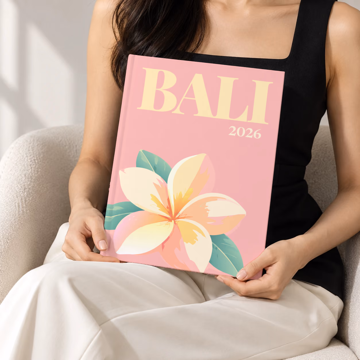 Bali hardcover photo book with the illustrated cover of a white and yellow frangipani flower on pink, held upright in two hands by a woman in a black top, sitting in a cream boucle armchair