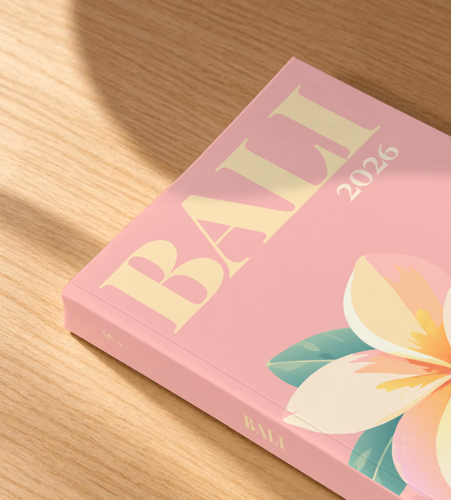 Bali softcover photo book with the illustrated cover of a white and yellow frangipani flower on pink, lying on an oak desk in dappled afternoon sunlight