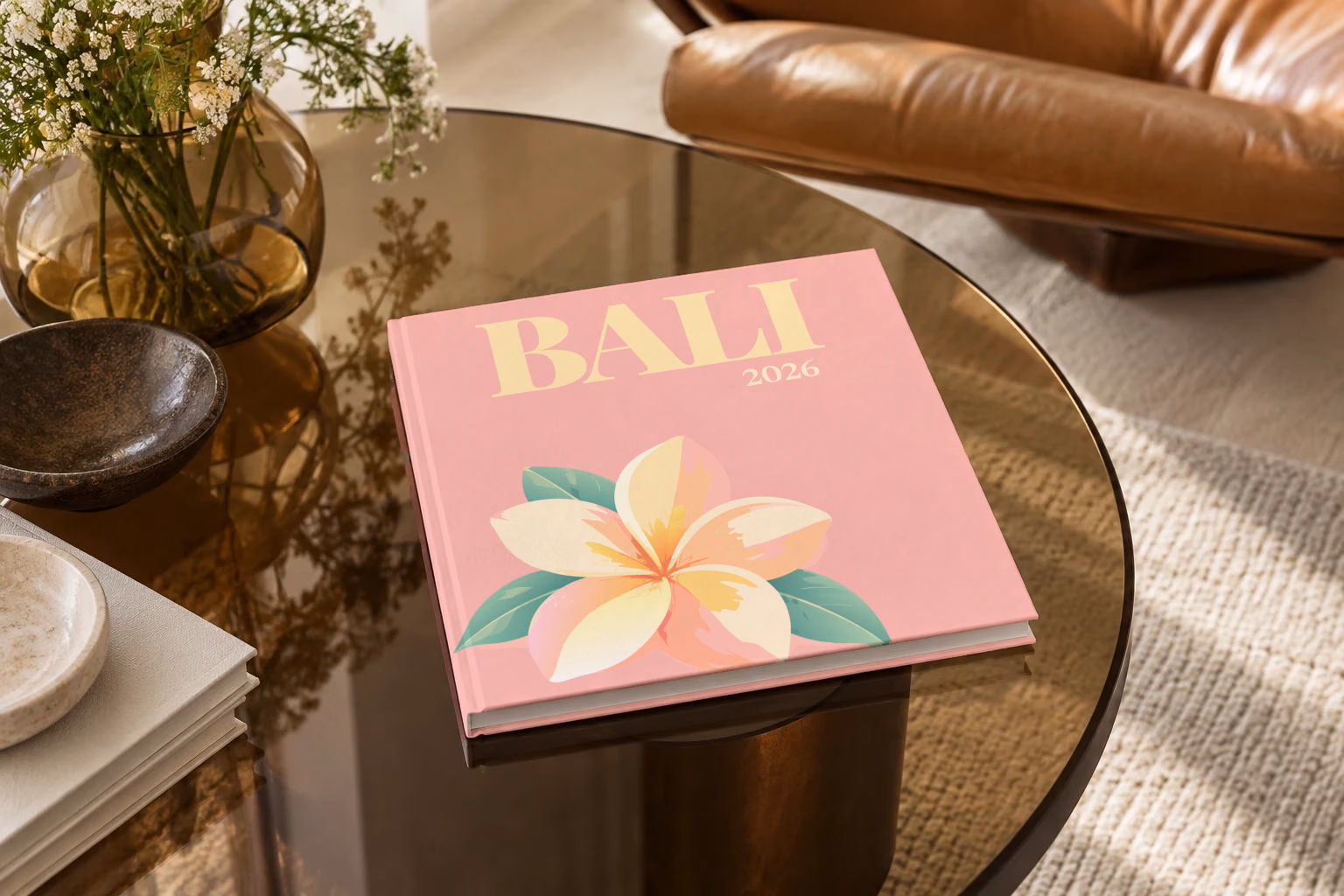 Bali square hardcover photo book with the illustrated cover of a white and yellow frangipani flower on pink, on a smoked glass coffee table beside a leather armchair in warm afternoon light