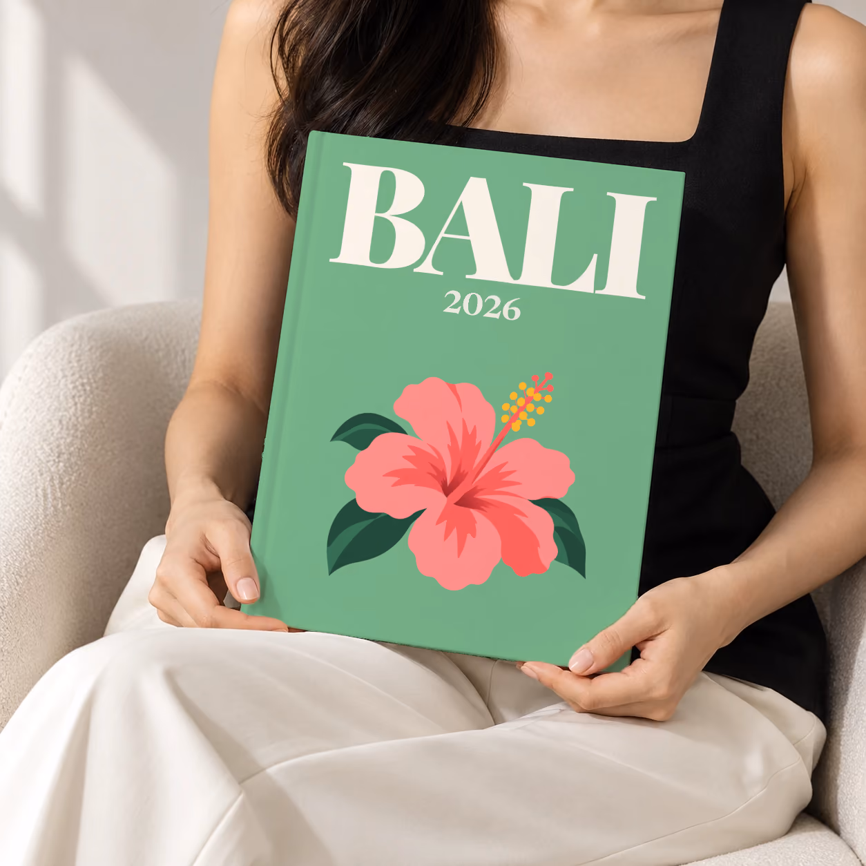 Bali hardcover photo book with the illustrated cover of a pink hibiscus flower on sage green, held upright in two hands by a woman in a black top, sitting in a cream boucle armchair