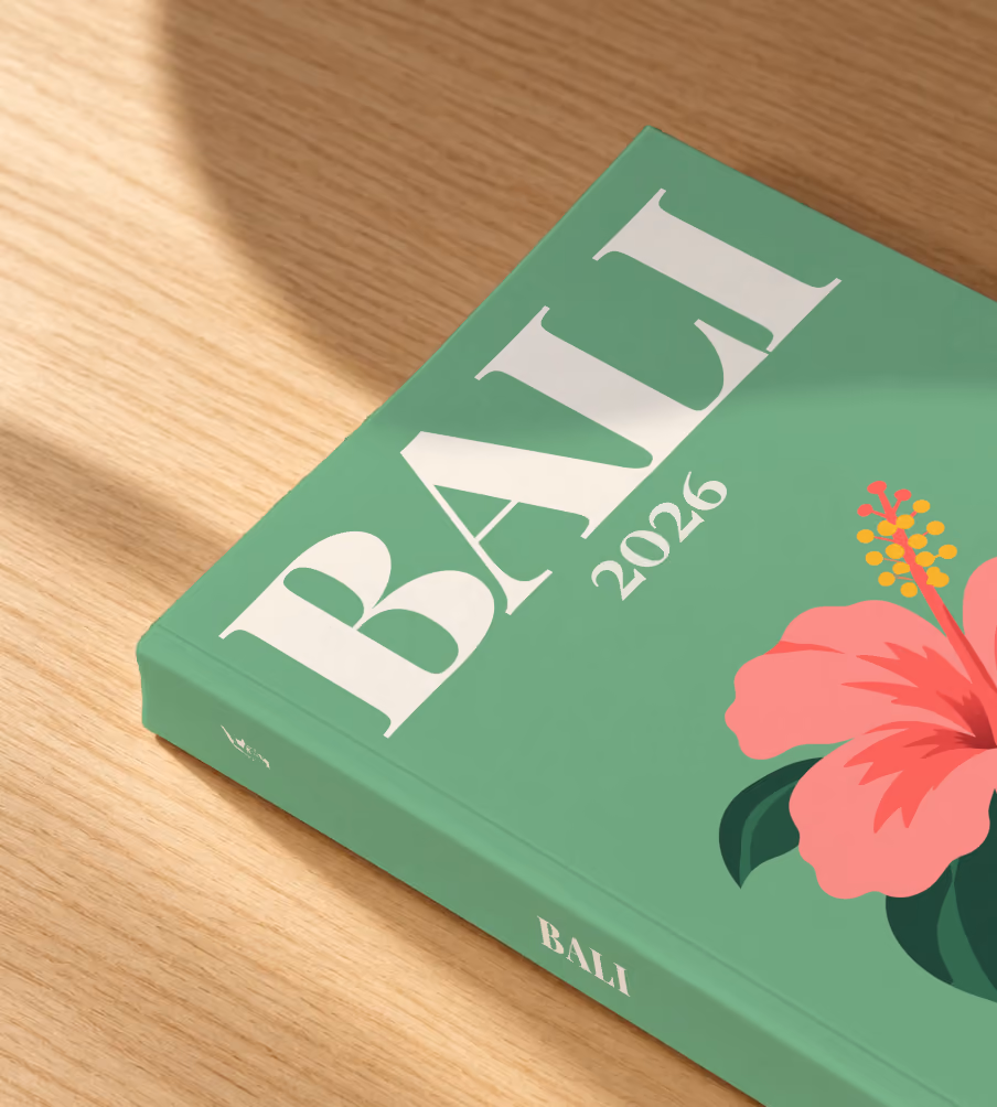 Bali softcover photo book with the illustrated cover of a pink hibiscus flower on sage green, lying on an oak desk in dappled afternoon sunlight
