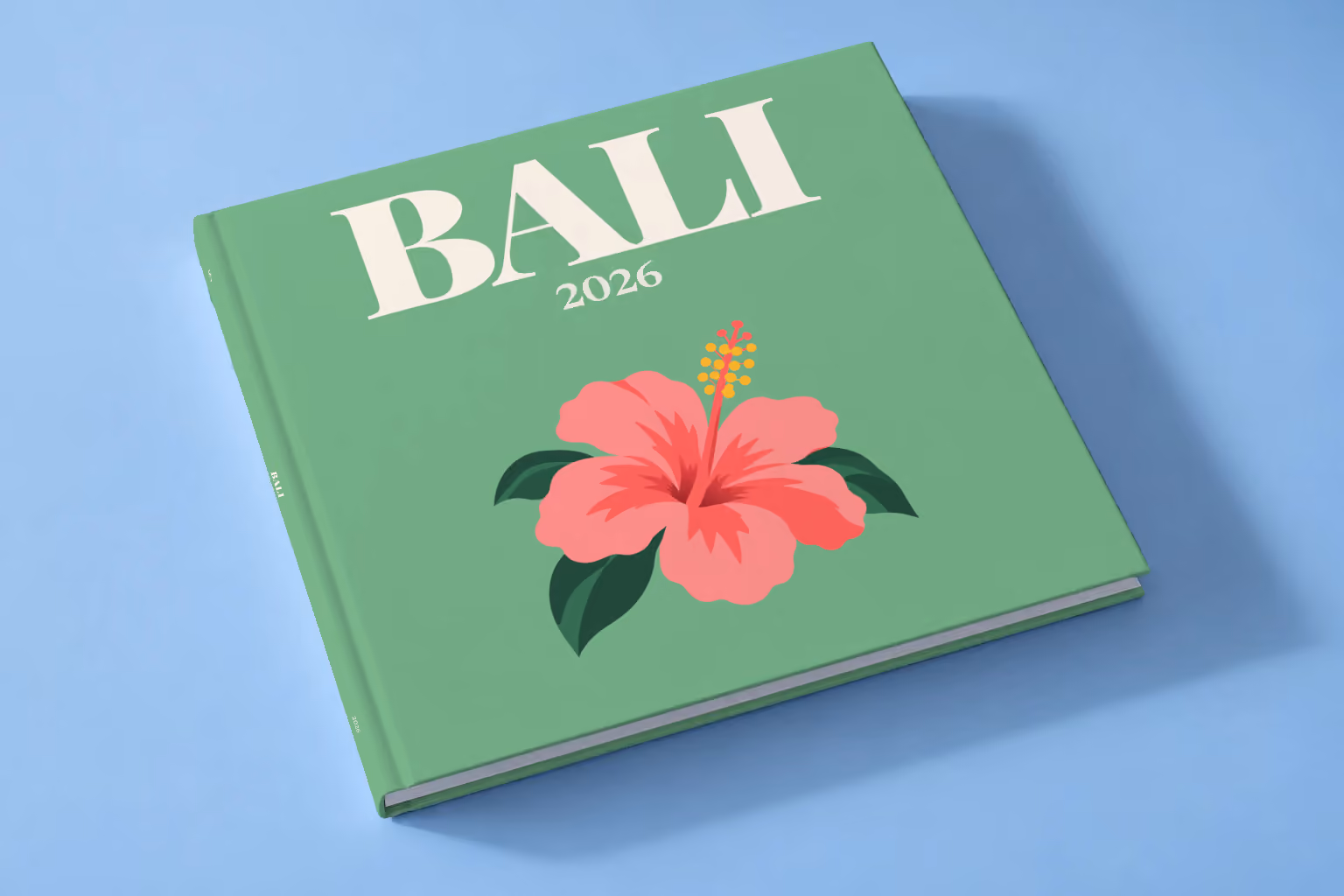 Bali square hardcover photo book with the illustrated cover of a pink hibiscus flower on sage green, lying on a pale blue studio backdrop