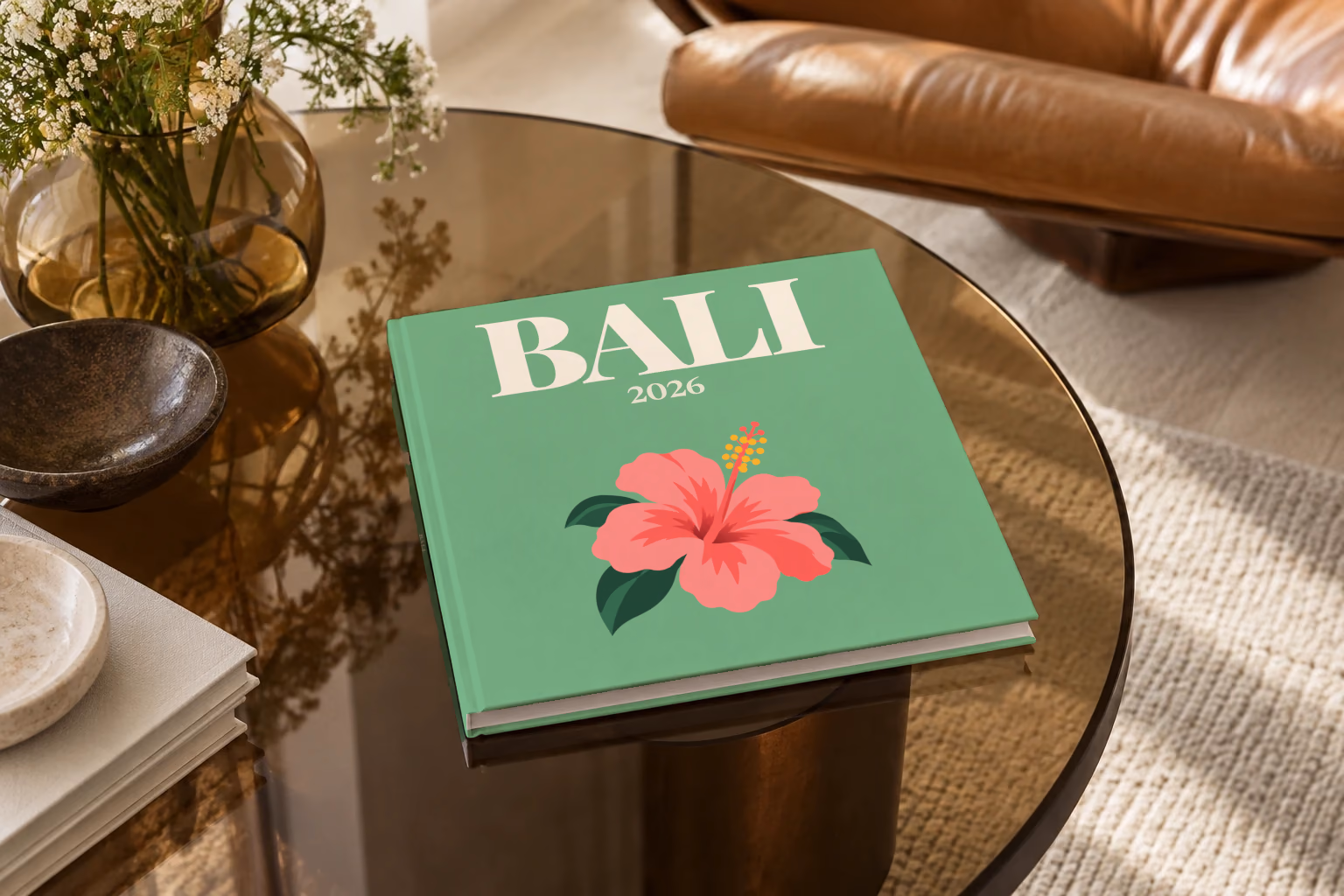 Bali square hardcover photo book with the illustrated cover of a pink hibiscus flower on sage green, on a smoked glass coffee table beside a leather armchair in warm afternoon light