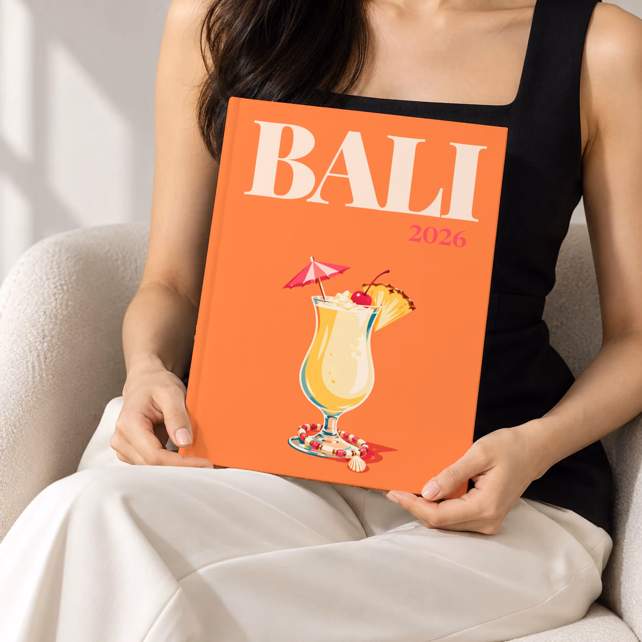 Bali hardcover photo book with the illustrated cover of a piña colada with a paper umbrella, a cherry and pineapple on orange, held upright in two hands by a woman in a black top, sitting in a cream boucle armchair
