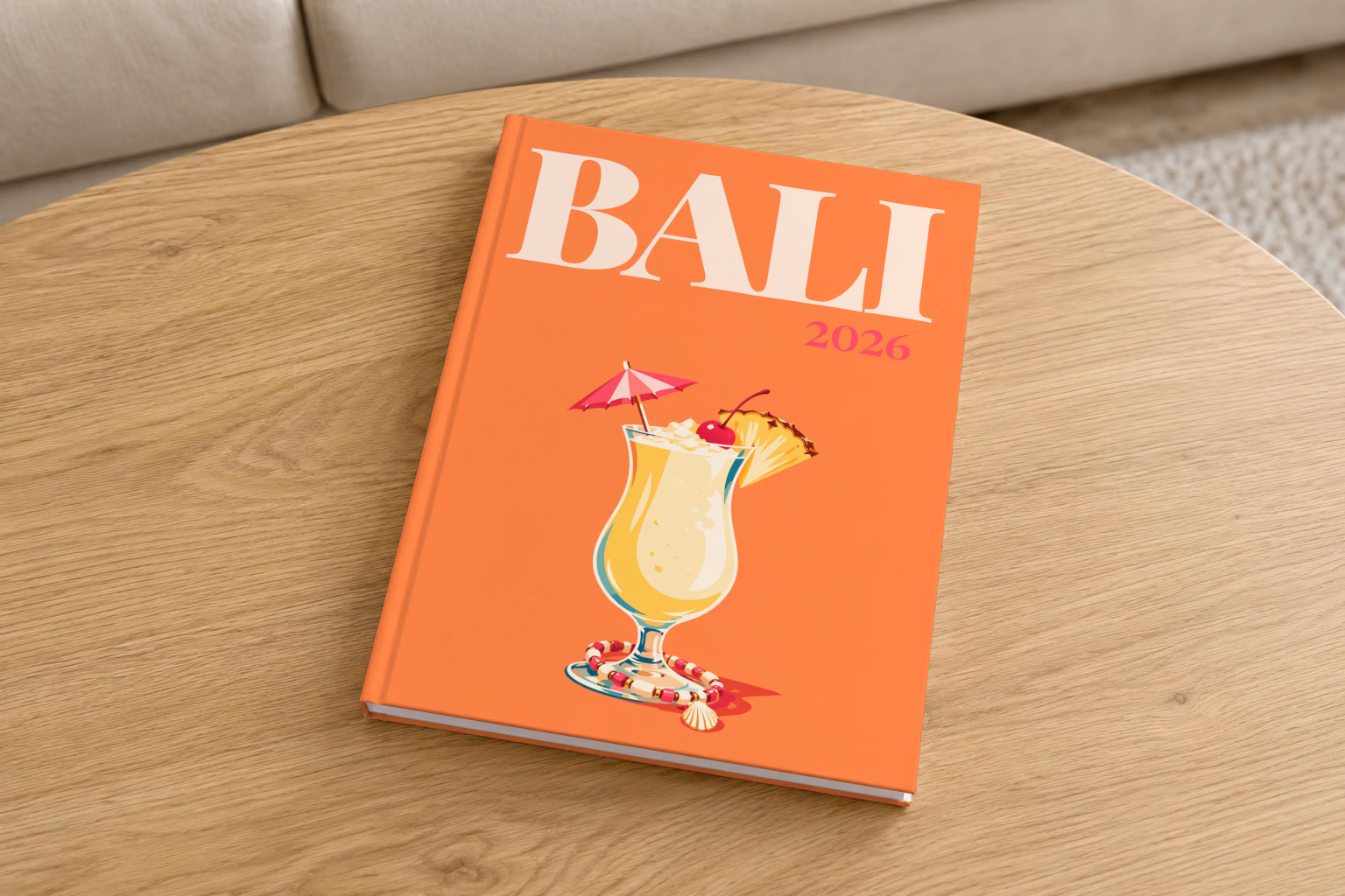 Bali hardcover photo book with the illustrated cover of a piña colada with a paper umbrella, a cherry and pineapple on orange, on a round oak coffee table in a bright living room