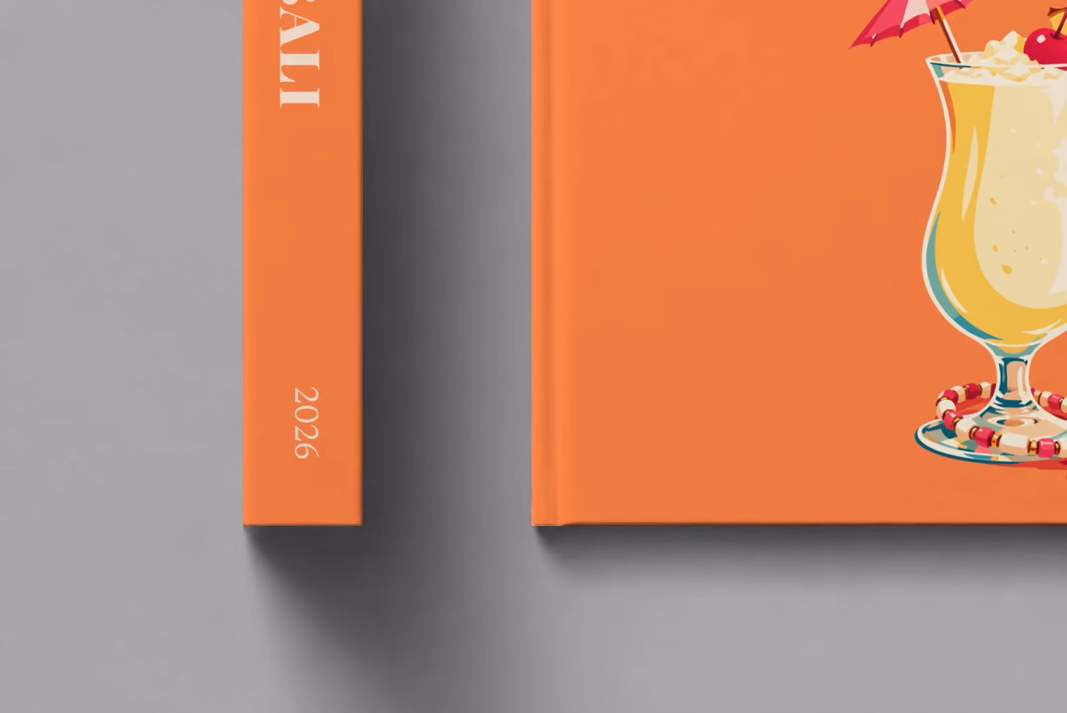 Bali hardcover photo book with the illustrated cover of a piña colada with a paper umbrella, a cherry and pineapple on orange, shown close up beside its spine on a pale grey studio surface