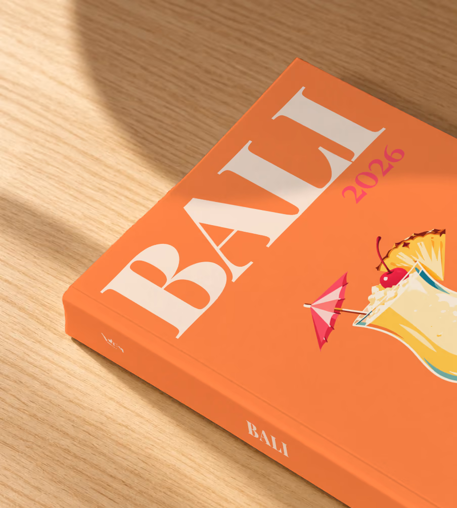 Bali softcover photo book with the illustrated cover of a piña colada with a paper umbrella, a cherry and pineapple on orange, lying on an oak desk in dappled afternoon sunlight