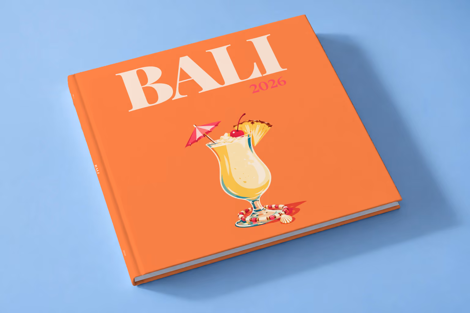 Bali square hardcover photo book with the illustrated cover of a piña colada with a paper umbrella, a cherry and pineapple on orange, lying on a pale blue studio backdrop