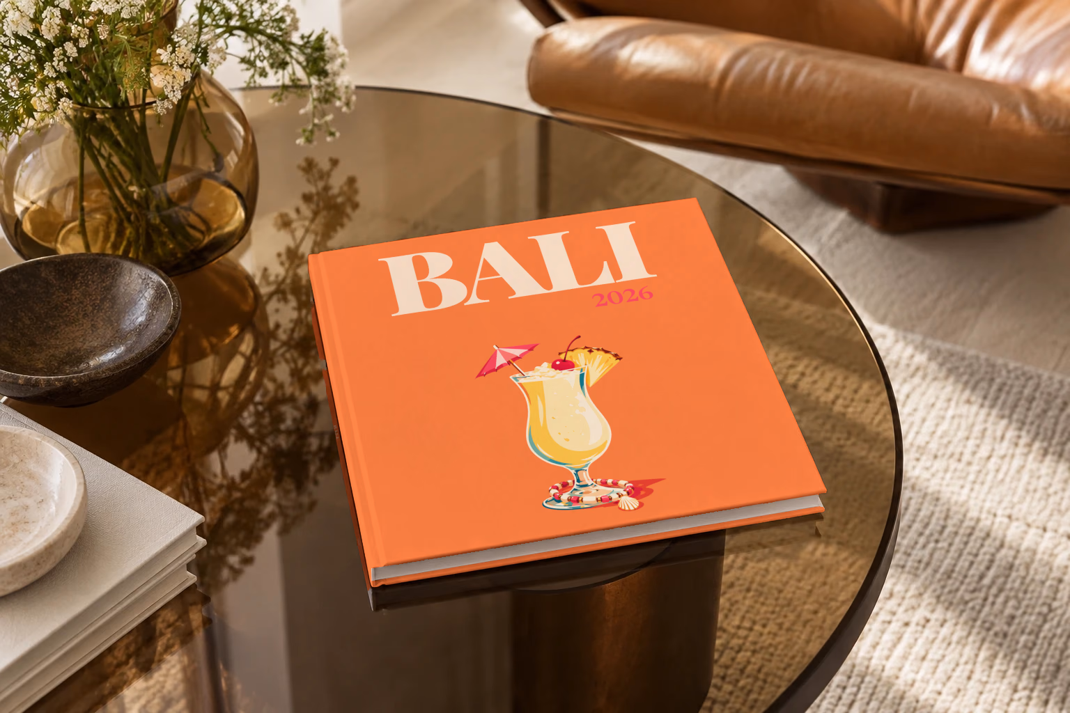 Bali square hardcover photo book with the illustrated cover of a piña colada with a paper umbrella, a cherry and pineapple on orange, on a smoked glass coffee table beside a leather armchair in warm afternoon light