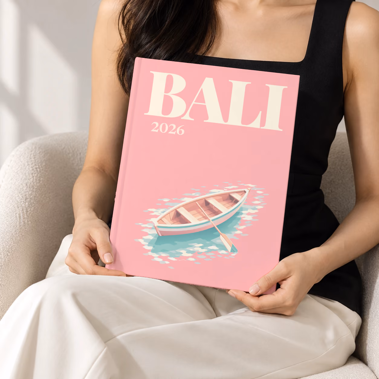 Bali hardcover photo book with the illustrated cover of a wooden rowboat on clear water on pink, held upright in two hands by a woman in a black top, sitting in a cream boucle armchair