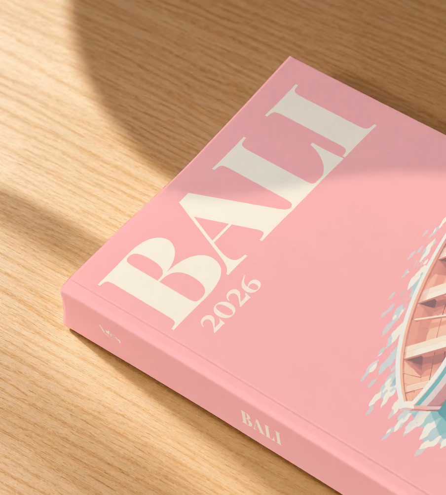 Bali softcover photo book with the illustrated cover of a wooden rowboat on clear water on pink, lying on an oak desk in dappled afternoon sunlight