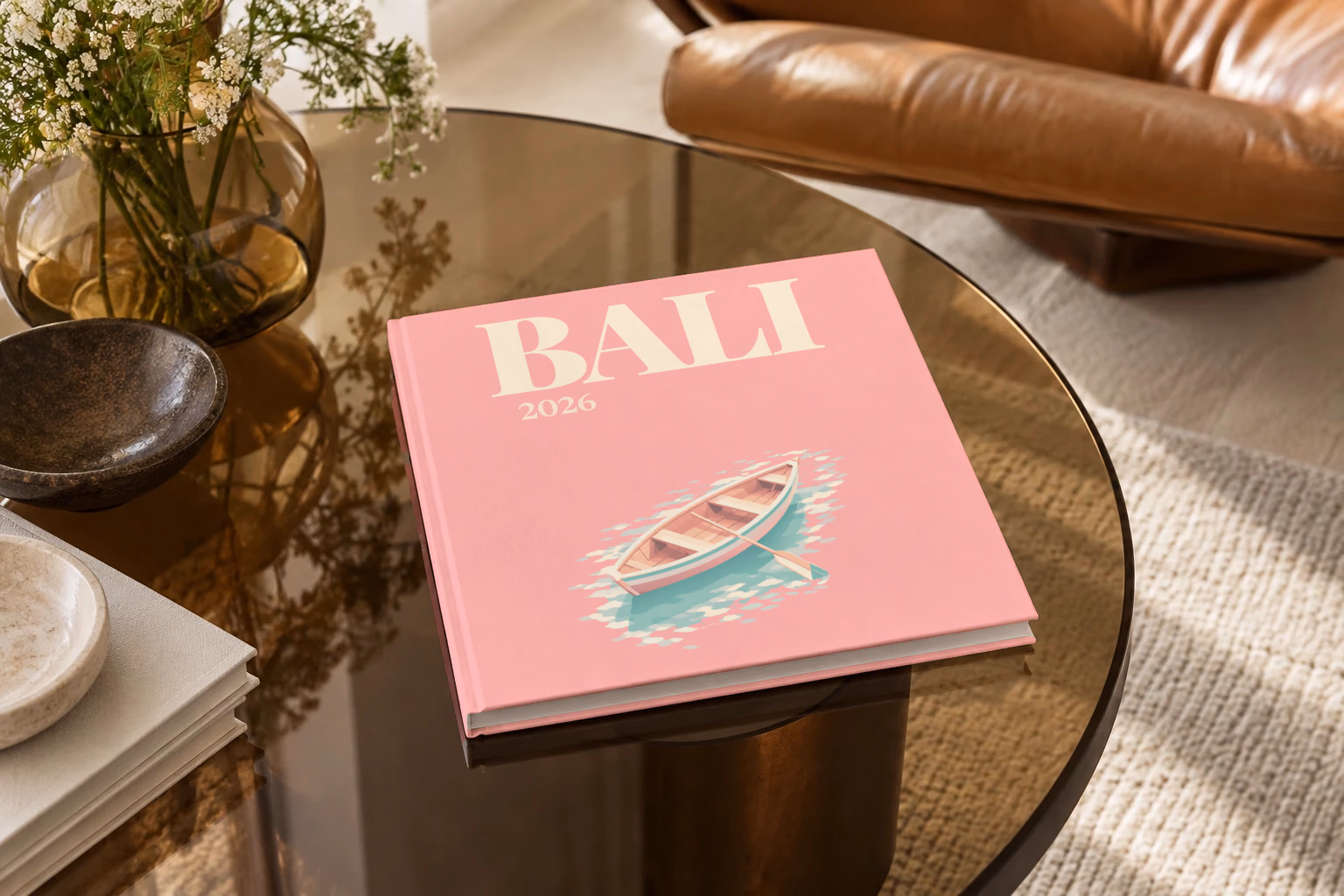 Bali square hardcover photo book with the illustrated cover of a wooden rowboat on clear water on pink, on a smoked glass coffee table beside a leather armchair in warm afternoon light