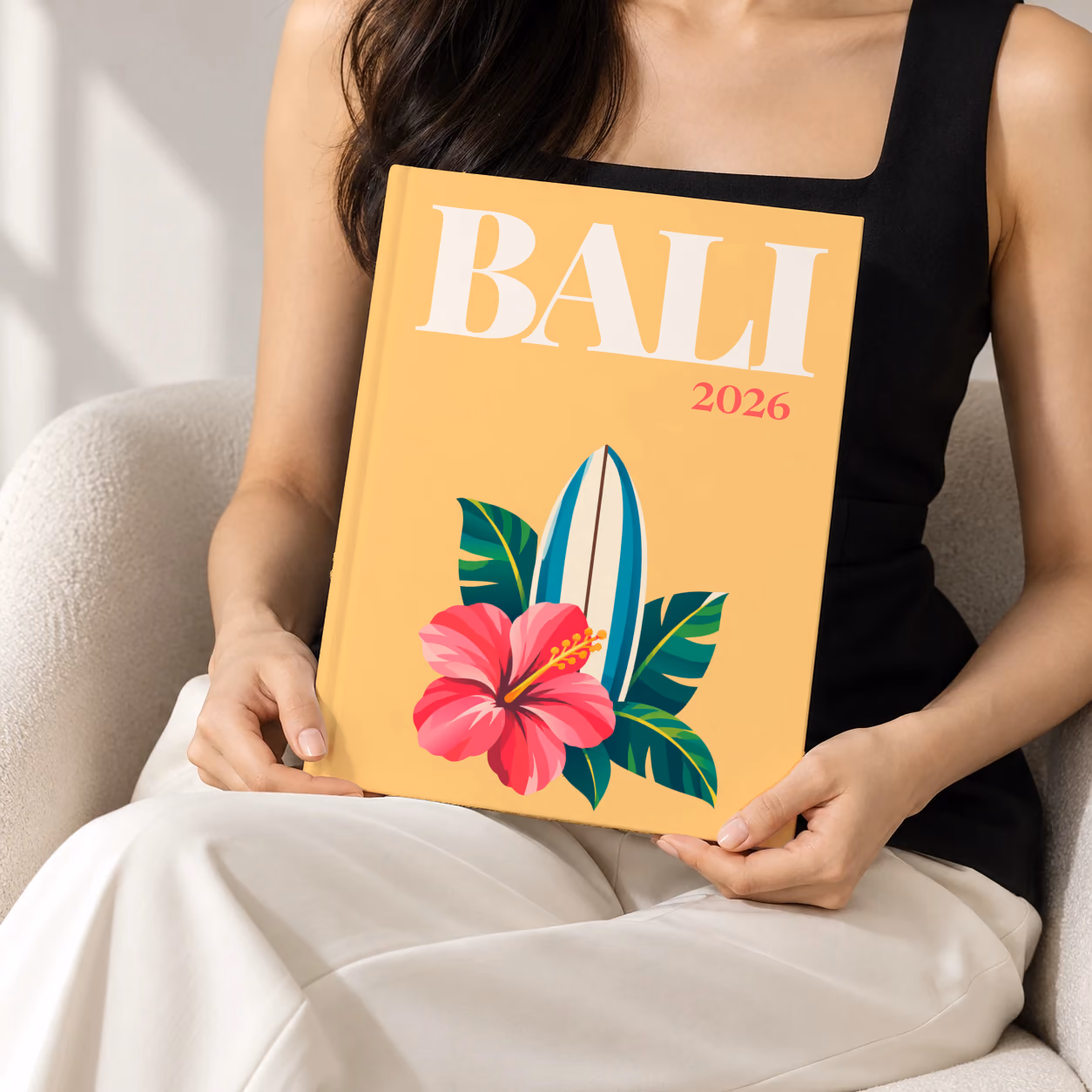 Bali hardcover photo book with the illustrated cover of a surfboard with a pink hibiscus and palm leaves on apricot, held upright in two hands by a woman in a black top, sitting in a cream boucle armchair