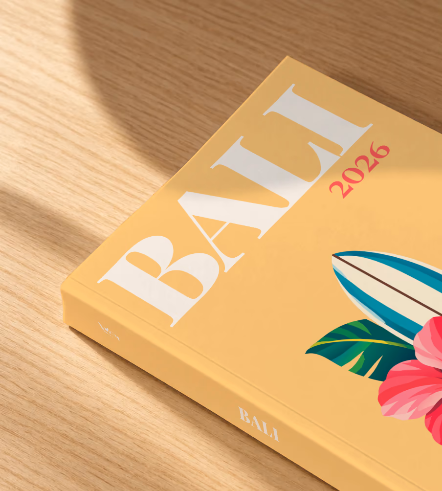 Bali softcover photo book with the illustrated cover of a surfboard with a pink hibiscus and palm leaves on apricot, lying on an oak desk in dappled afternoon sunlight