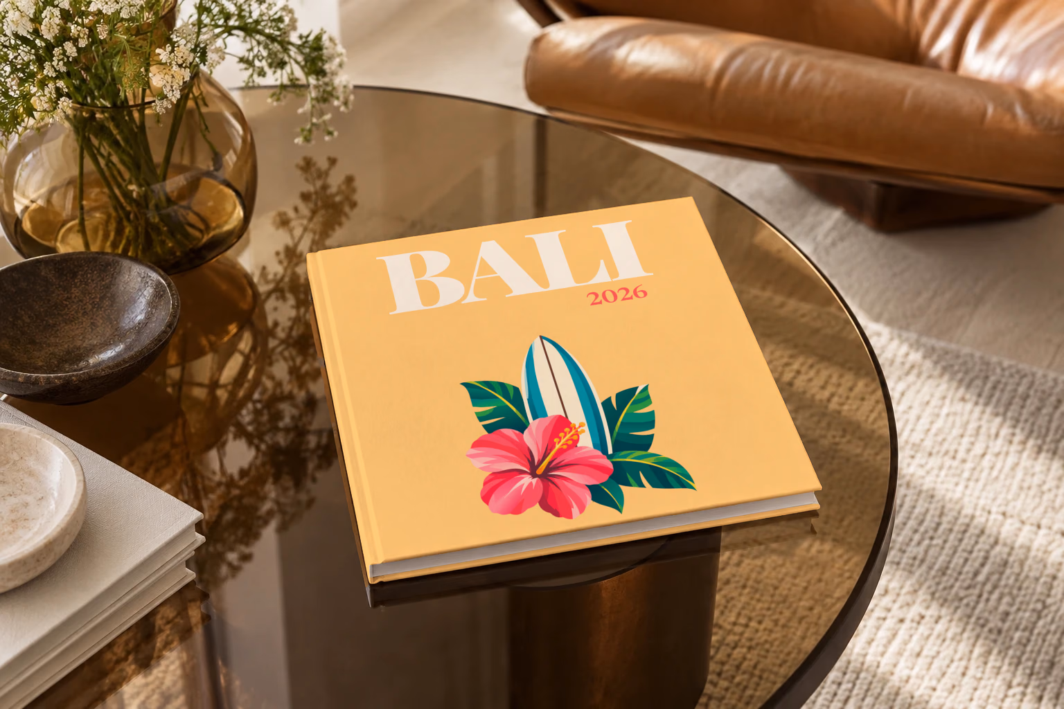 Bali square hardcover photo book with the illustrated cover of a surfboard with a pink hibiscus and palm leaves on apricot, on a smoked glass coffee table beside a leather armchair in warm afternoon light