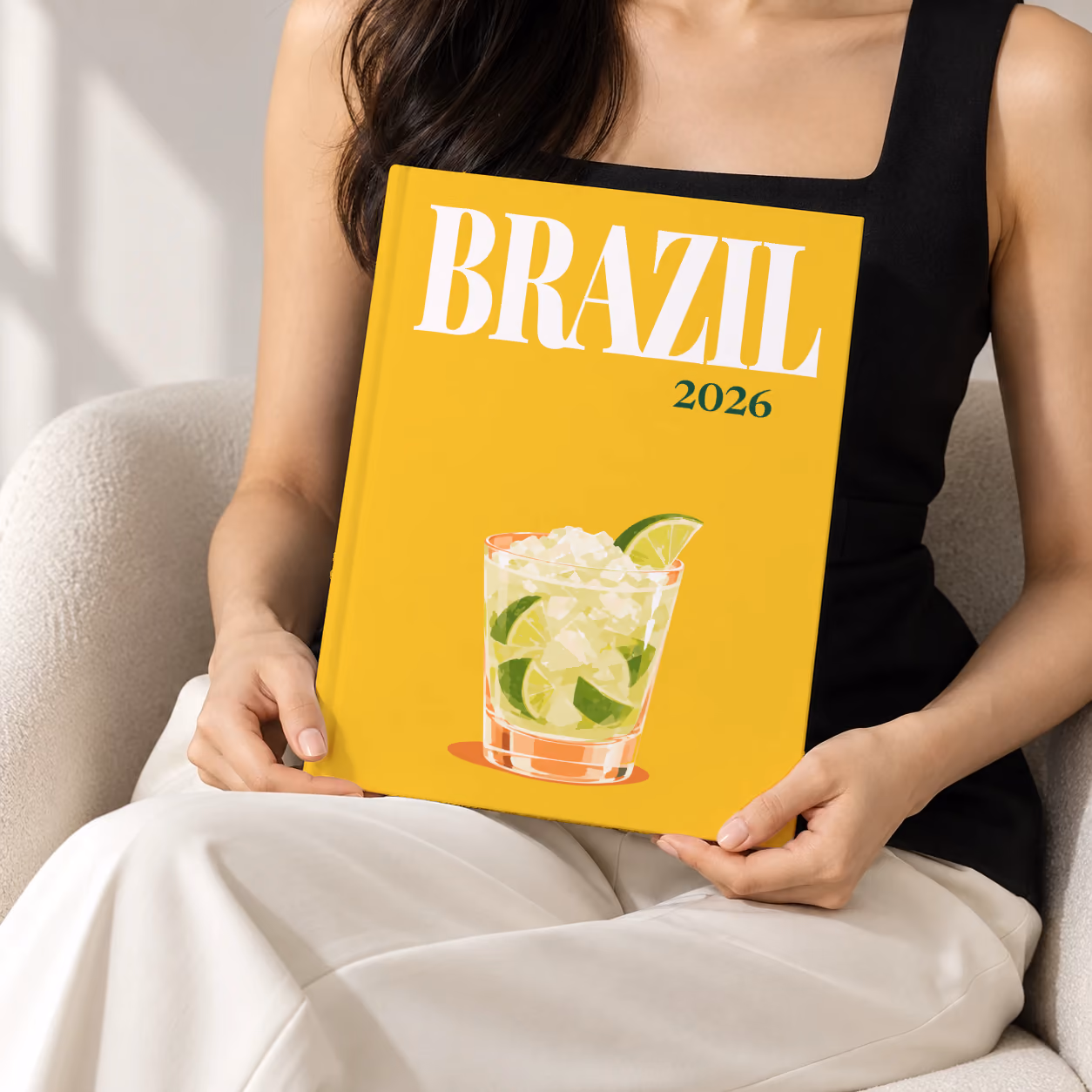 Brazil hardcover photo book with the illustrated cover of a caipirinha with crushed ice and lime on golden yellow, held upright in two hands by a woman in a black top, sitting in a cream boucle armchair