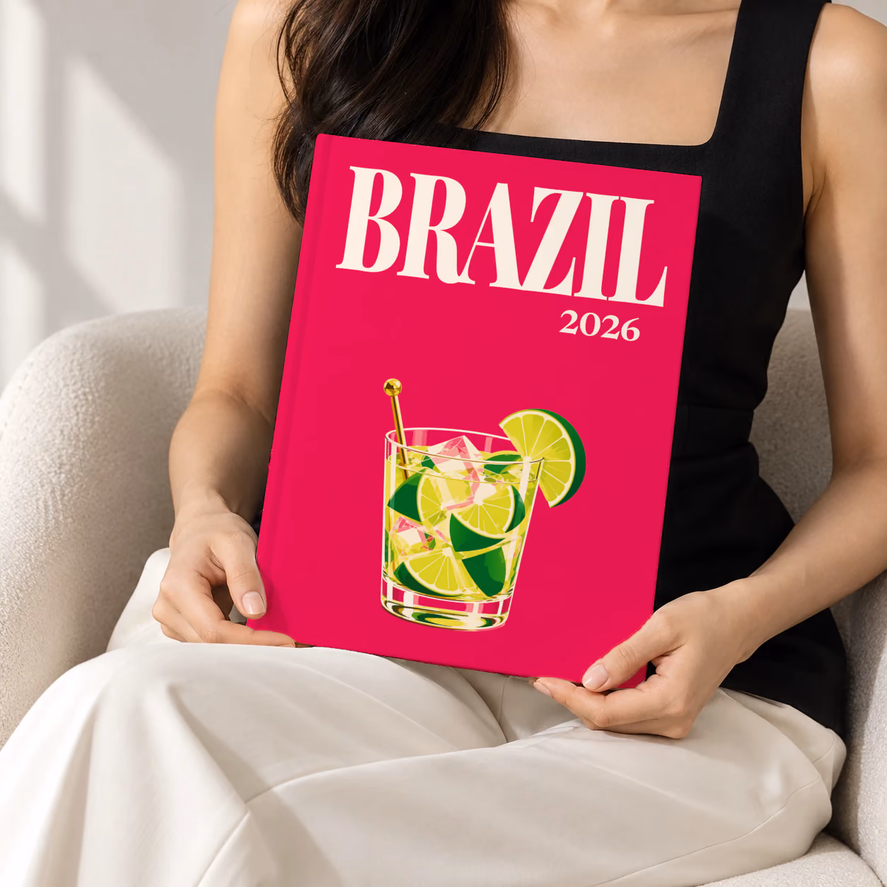 Brazil hardcover photo book with the illustrated cover of a caipirinha with lime wedges on magenta, held upright in two hands by a woman in a black top, sitting in a cream boucle armchair