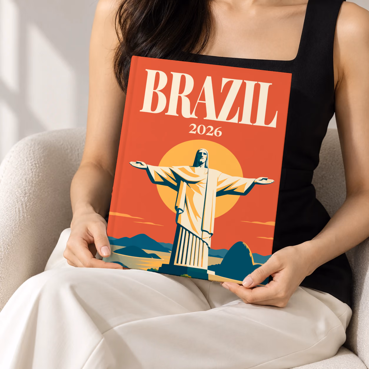 Brazil hardcover photo book with the illustrated cover of Christ the Redeemer above Rio de Janeiro's bay at sunset on red-orange, held upright in two hands by a woman in a black top, sitting in a cream boucle armchair