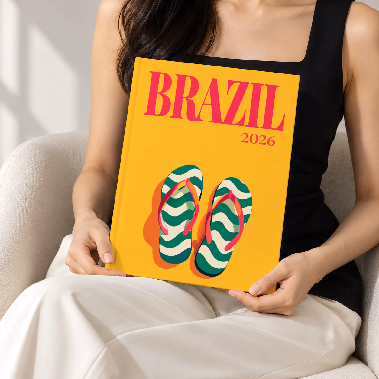 Brazil hardcover photo book with the illustrated cover of a pair of flip-flops in Copacabana's wave pattern on yellow, held upright in two hands by a woman in a black top, sitting in a cream boucle armchair