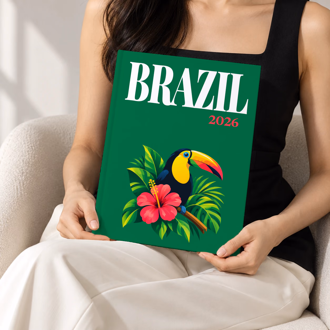 Brazil hardcover photo book with the illustrated cover of a toucan among tropical leaves and a pink hibiscus on emerald green, held upright in two hands by a woman in a black top, sitting in a cream boucle armchair