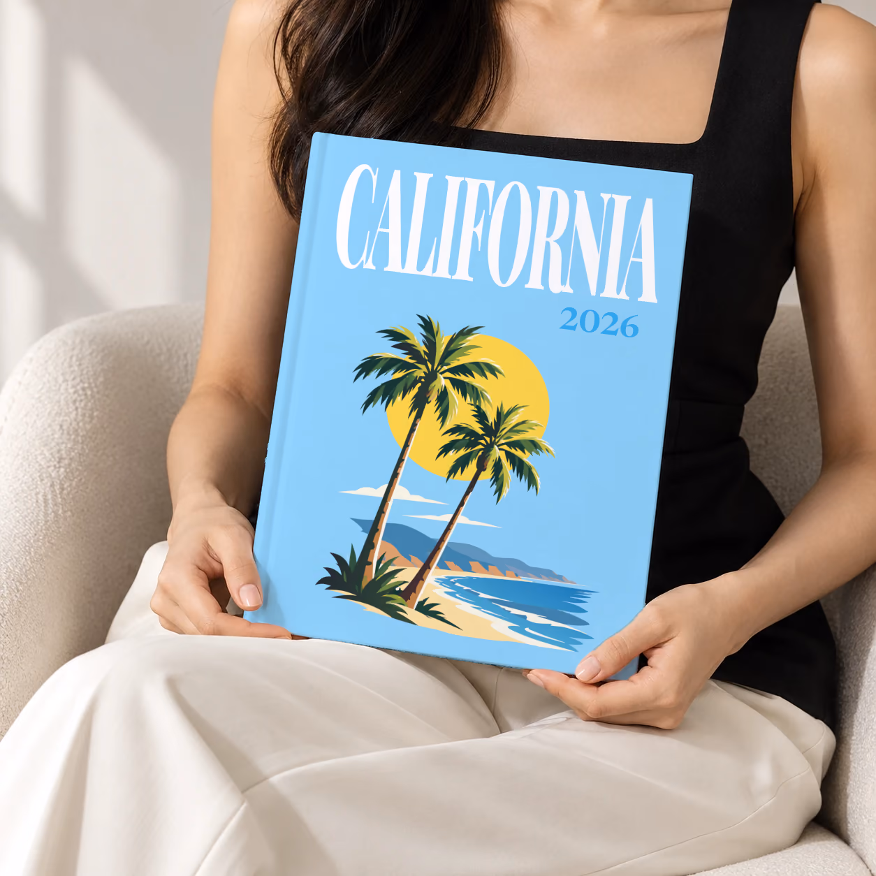 California hardcover photo book with the illustrated cover of palm trees and a beach under a yellow sun on sky blue, held upright in two hands by a woman in a black top, sitting in a cream boucle armchair