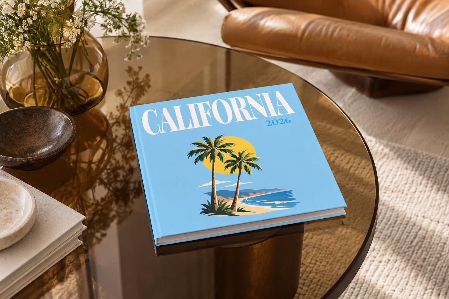 California square hardcover photo book with the illustrated cover of palm trees and a beach under a yellow sun on sky blue, on a smoked glass coffee table beside a leather armchair in warm afternoon light
