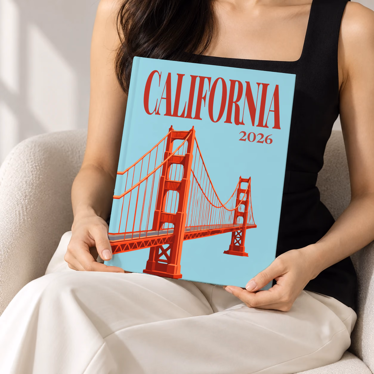 California hardcover photo book with the illustrated cover of the Golden Gate Bridge on pale aqua, held upright in two hands by a woman in a black top, sitting in a cream boucle armchair