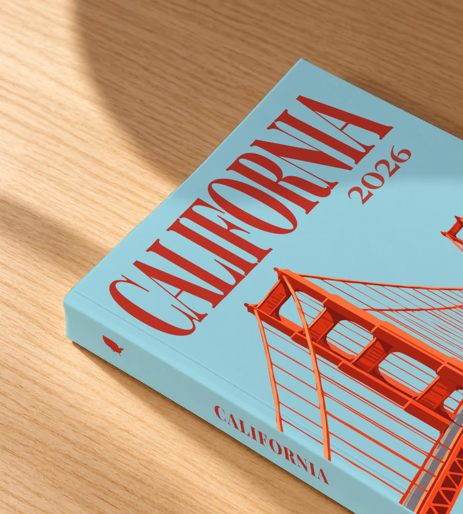 California softcover photo book with the illustrated cover of the Golden Gate Bridge on pale aqua, lying on an oak desk in dappled afternoon sunlight