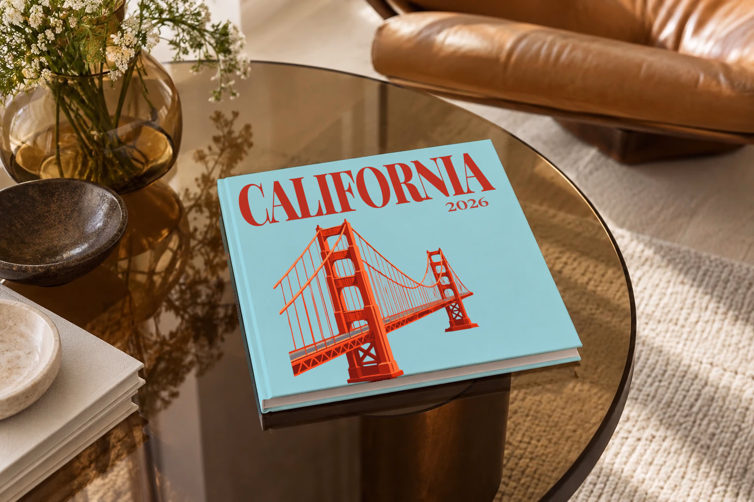 California square hardcover photo book with the illustrated cover of the Golden Gate Bridge on pale aqua, on a smoked glass coffee table beside a leather armchair in warm afternoon light